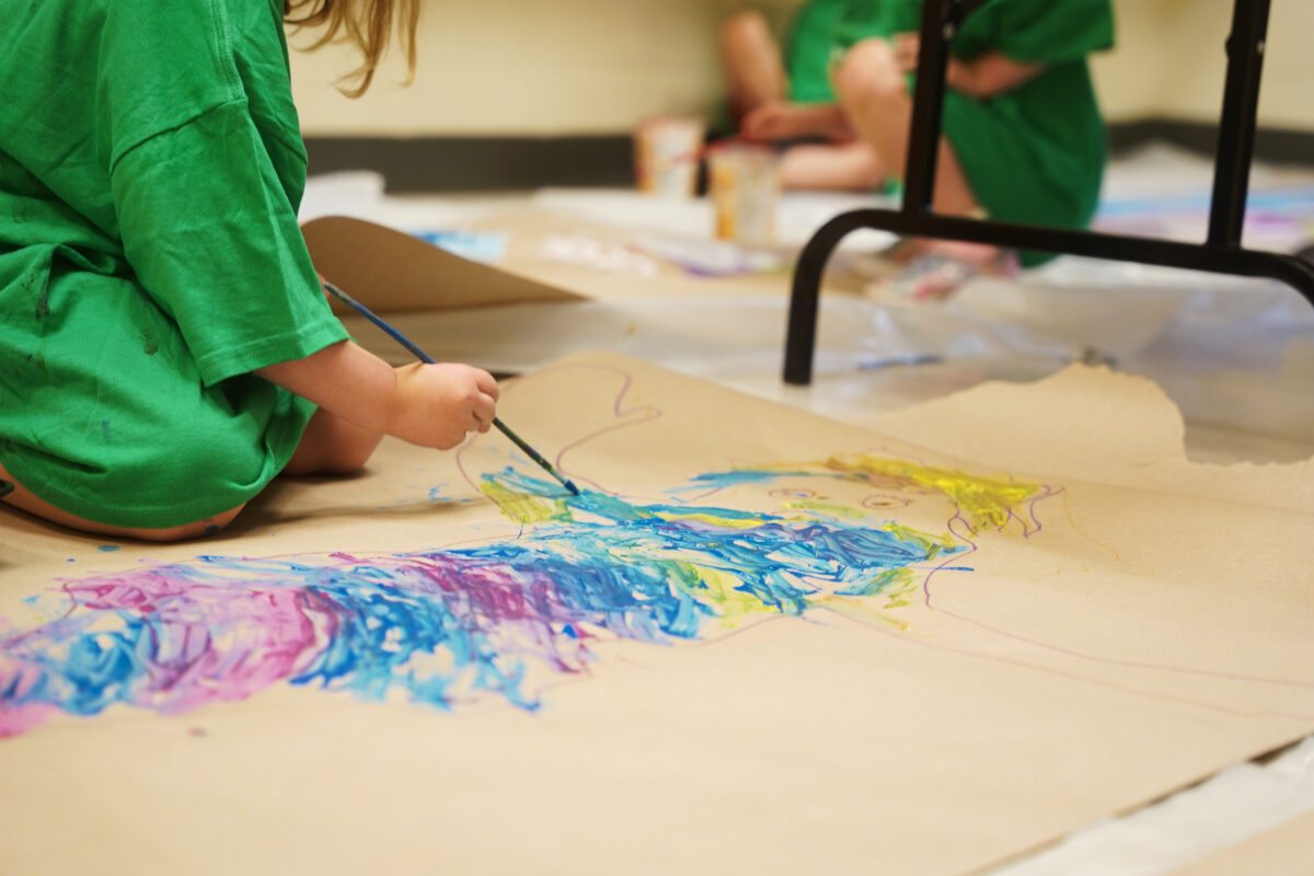 Image of a young person in a green paint shirt painting a large piece of paper with blues, purples and yellow
