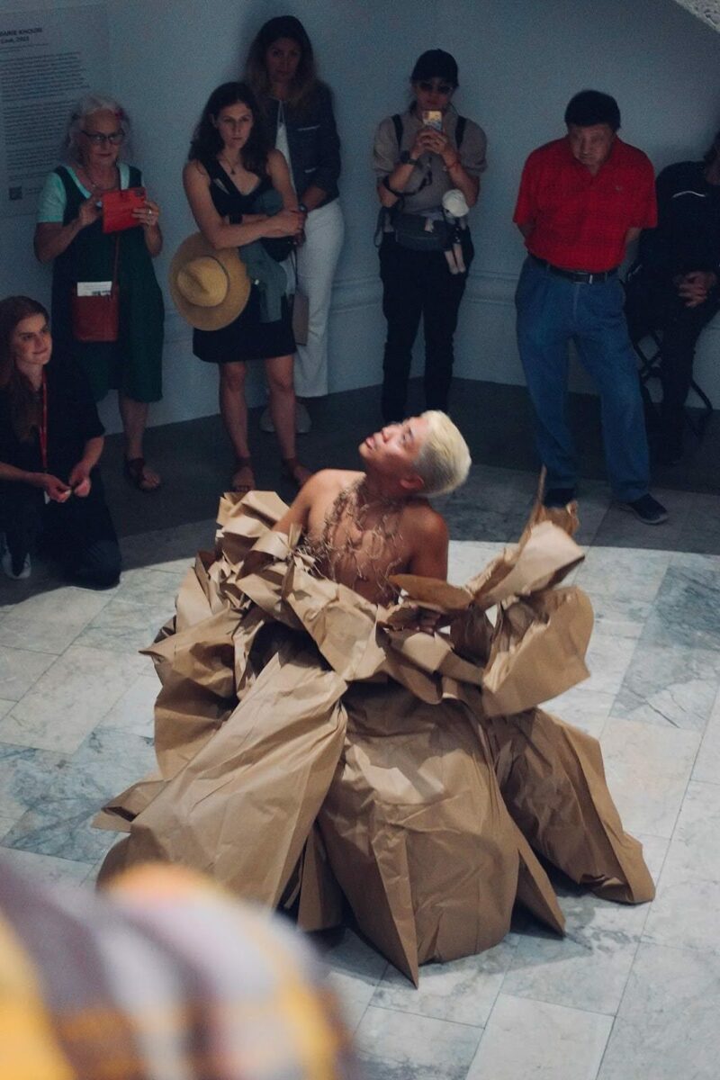 A dancer stands in the centre of a grey tiled floor dressed in a voluminous brown paper dress. Dancer is looking up towards their right. Seven audience members are seen standing behind the dancer.