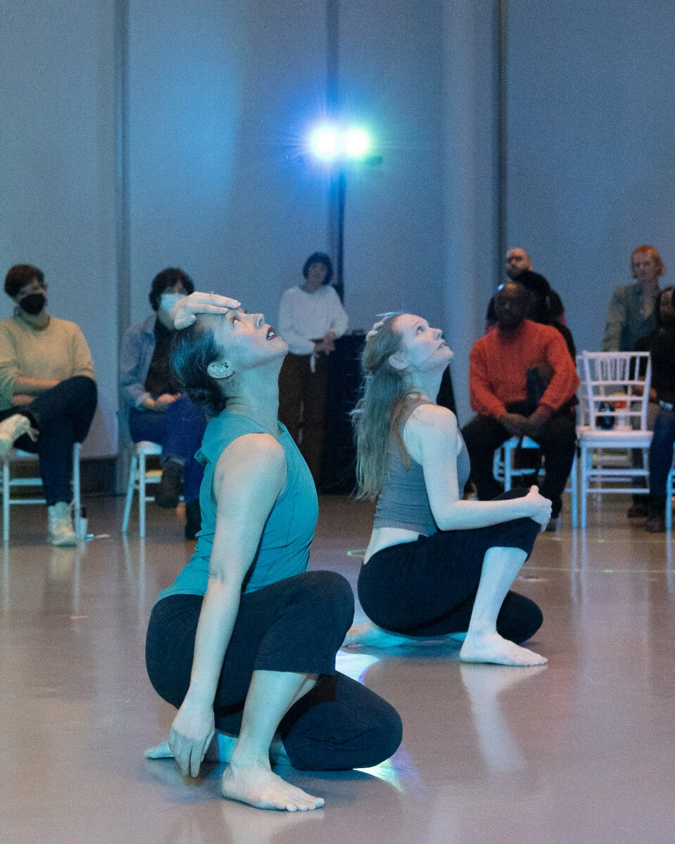 Two dancers squat on on knee. Each has one hand placed on their forehead and are looking up.