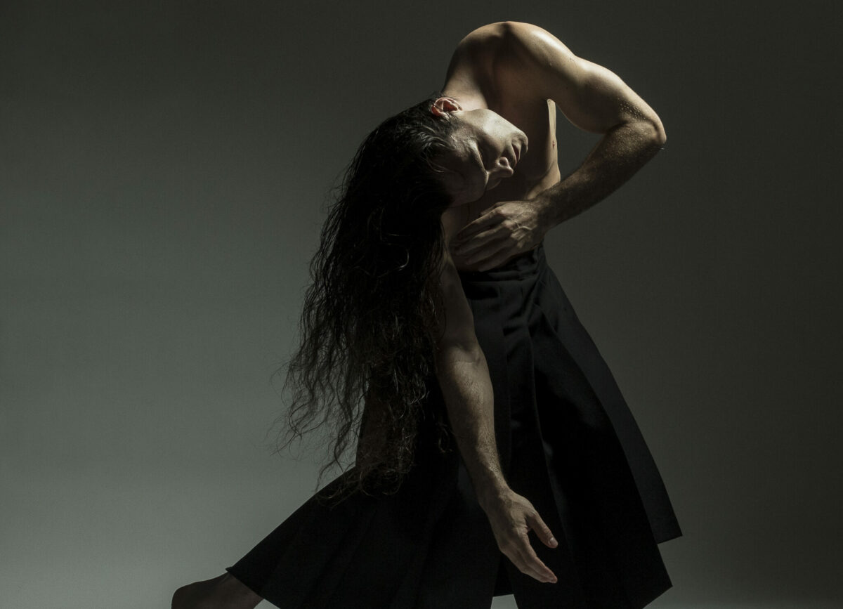 Dancer Jera Wolfe who has long brown hair, wearing drapey black pants is leaning towards one leg with right arm hanging and left arm bent touching right shoulder. The background is a grey gradient.