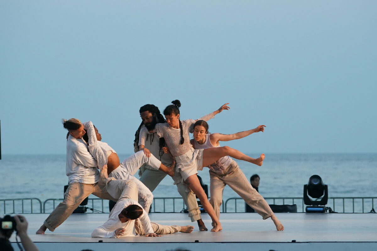 Six dancers dressed in white and beige in an interconnected formation. One dancer is on the floor at the front. Three dancers at the back are standing and supporting the weight of two off-balance dancers. They are on an outdoor stage, the background is a blue sky and blue water.